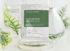 Embalage do novo Booster para tratamento de sobrancelhas em um fundo remetendo a um laboratório, com detalhes brancos com folhas verdes